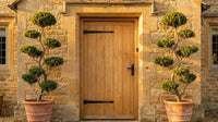 Showcase · Two cloud-pruned evergreens in terracotta pots flank an oak door in warm honey-coloured stone.