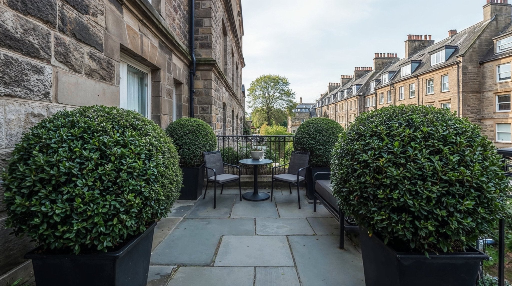 Formal balcony terrace featuring a matched pair of Ilex ball topiary in black planters, framing a small seating area with chairs and table, set against traditional UK stone architecture and a residential streetscape.